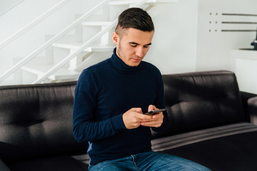 Attractive young man using mobile phone, texting, sitting on black couch. Dressed in jeans and sweater. Modern home interior.