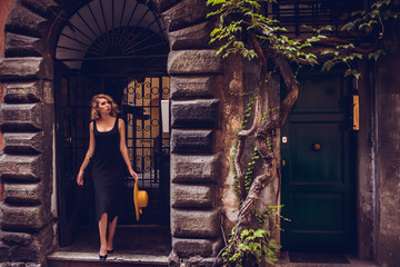 Vogue style elegant portrait of beautiful fashion woman wavy shine blonde long hair. Model in yellow summer hat black dress and luxury accessories with bright makeup at street of Trastevere Rome Italy