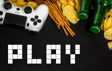 Modern white gamepad among chips, crackers and bottles of beer on a black. Game concept.