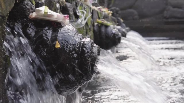 Close-up of holy spring water stream pouring into pool and traditional Balinese offerings Canang sari made of leaves and flowers in Pura Tirta Empul Temple on Bali, Indonesia. Camera stays still.