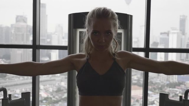 Portrait of beautiful blonde woman at the gym exercising on a training machine. Gym with big windows. Healthy and active lifestyle. Slow motion shot.