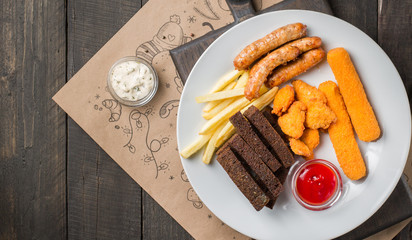 Fried beer appetizer. French fries, chicken nuggets, cheese fingers, sausage, tomato sauce and bread crumbs on white plate on a dark wooden background. Street fast food. Flat lay, top view.