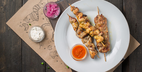 Assorted delicious grilled meat with sauces on white plate on a dark wooden background.  BBQ plate with different kinds of meat. Street fast food. Flat lay, top view.
