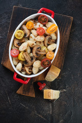 Bowl of potato gnocchi with roasted mushrooms and fresh sliced tomatoes on a dark brown stone background, vertical shot, above view