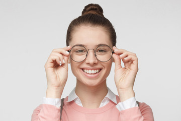 Closeup of attractive young woman isolated on gray background, wearing big round spectacles with thin black frame, touching temples with fingers, smiling