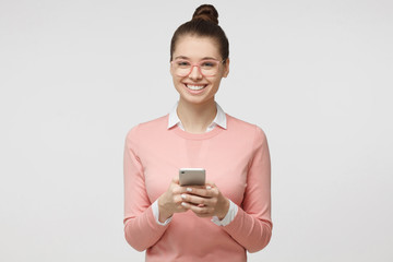 Indoor shot of good looking young woman isolated on gray background looking at smartphone, smiling openly while holding smartphone in both hands