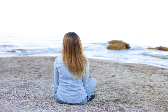 Happy girl sits with his back to the camera and enjoying pleasant pastime, sitting on beach and looking toward endless blue sea on pleasant evening. Young European-looking woman with blond long  hai