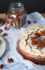 Angle view of cake with sliced banana, walnut and powdered sugar and glass jar full of nuts. Cozy still life in light tones on background with checked kitchen towel.
