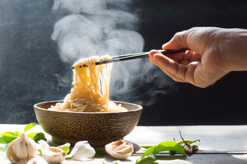Hand uses chopsticks to pickup tasty noodles with steam and smoke in bowl on wooden background,...