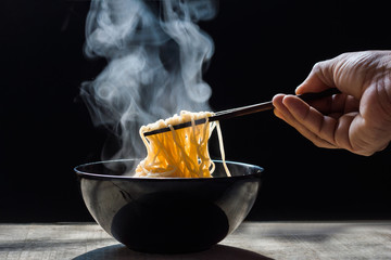 Die Hand verwendet Essstäbchen, um leckere Nudeln mit Dampf und Rauch in der Schüssel auf Holzhintergrund aufzunehmen, selektiver Fokus. Draufsicht, asiatische Mahlzeit auf einem Tisch, Junk-Food-Konzept