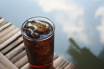 Glass of soft drink or cola with ice cubes on bamboo raft in the lake background and reflection of sky in water top view