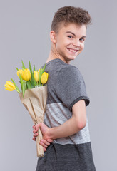 Teen boy hiding bunch of flowers behind itself, on gray background. Child with bouquet of yellow tulips as a gift. Happy mothers or Valentines day