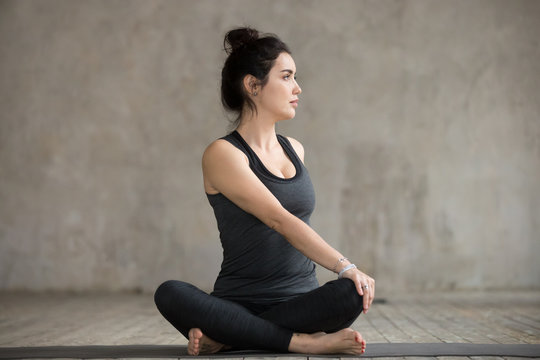 Young sporty woman practicing yoga, doing Revolved Easy exercise, Parivrtta Sukhasana pose, working out, wearing sportswear, black pants and top, indoor full length, gray wall, yoga studio