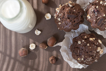 Three chocolate cupcakes, a small bottle of milk and a few hazelnuts on the brown background with the shadow of the shutters.