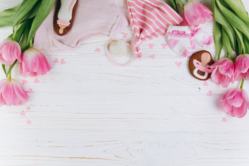 A composition for newborns on a wooden white background with clothes, pink tulips, hearts and a cookies, copy space and flat lay. It's a girl.