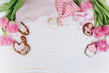A composition for newborns on a wooden white background with clothes, pink tulips, hearts and a cookies, copy space and flat lay. It's a girl.