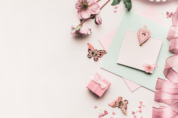 Mock up of feminine holidays greeting card in pale pastel color with flowers, gift box,ribbon and  heart on white desktop background, top view. Styled flat lay .