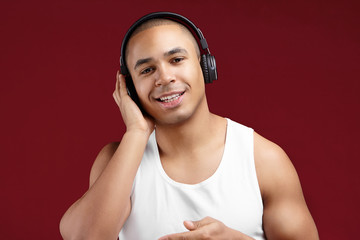 Cropped shot of handsome charismatic young dark skinned male DJ in white sleeveless t-shirt smiling joyfully, holding hand on his ear wearing wireless headphones while choosing tracks for party