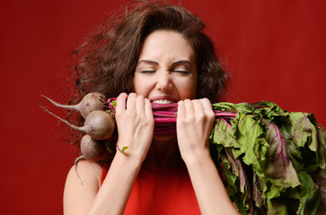 Young sport woman bite eat fresh beetroot with green leaves. Healthy eating concept on red background - obrazy, fototapety, plakaty