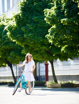 Beautiful female biker in white dress stopped for a moment looking in distance while riding her blue bike in city center with green trees on background. Pretty woman concentrated relax summer