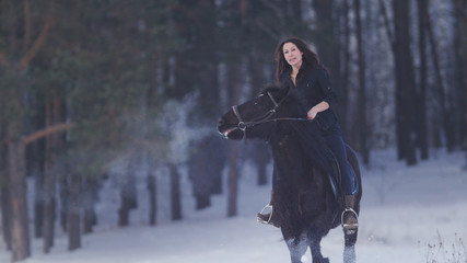 Beautiful longhaired woman riding a black horse through the snow in the forest, stallion prancing