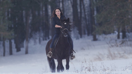 Attractive longhaired female rider riding a black horse through the drifts in the winter field