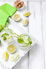 Fototapeta na wymiar Caipirinha, mojito cocktail with lime, brown sugar, ice and mint leaves in beautiful glasses, cut green citrus on white wooden background. Summer alcohol drink. Close up photography. Selective focus