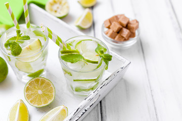 Caipirinha, mojito cocktail with lime, brown sugar, ice and mint leaves in beautiful glasses, cut green citrus on white wooden background. Summer alcohol drink. Close up photography. Selective focus