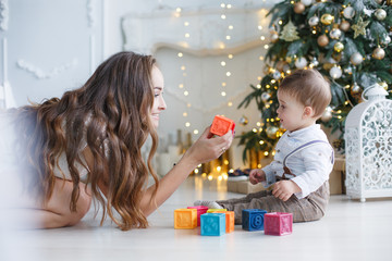 Obraz na płótnie Canvas A little boy in a white striped shirt and light brown pants with suspenders,plays with colored blocks with a beautiful young mother,a woman with long hair. Christmas party for mom and son