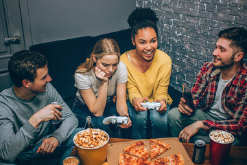 The blonde girl is frustrated because she has lost the game while her best friend has won it. Young men are just sitting with them. Gamers. Night party.