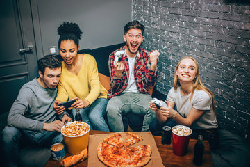 A small group of students sitting in the room. Two of them were playing game when suddenly the guy has won the battle and he is very happy about that. The other two are whatching film on the phone.