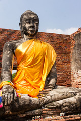 Old Buddha statue in festive safran clothes sitting in lotus position in ruined Wat in Ayuttaya of Thailand