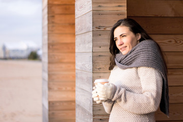 Young woman with natural face feeling cold wearing knitwear, holding hot coffee to go, looking aside