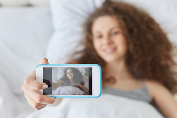 Female makes selfie with mobile phone, poses at camera, makes photo of herself after getting up in morning. Beautiful young woman uses modern gadget, shares photos in social networks online.