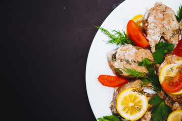 Baked pieces of pink salmon with vegetables, herbs and lemon on a white plate on a black background, close up.
