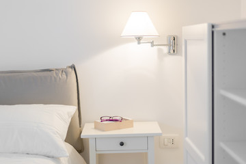 Detail of a bedroom. White interior. A lamp, a book and glasses on the nightstand. Relaxation concept