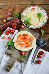 Ingredients for Pizza: basis dough, salami sausage, cherry tomatoes, grated cheese, tomato paste, herbs and arugula. Natural Wooden Background With Linen Napkin and Knife For Pizza