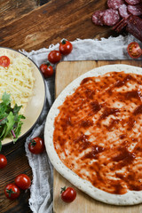 Ingredients for Pizza: basis dough, salami sausage, cherry tomatoes, grated cheese, tomato paste, herbs and arugula. Natural Wooden Background With Linen Napkin and Knife For Pizza