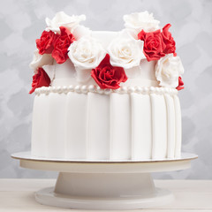 Two-tiered wedding cake decorated with red and white flowers from mastic on a white wooden table.