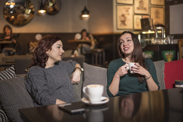 Two young beautiful girls sitting in urban cafe with coffee and talking.