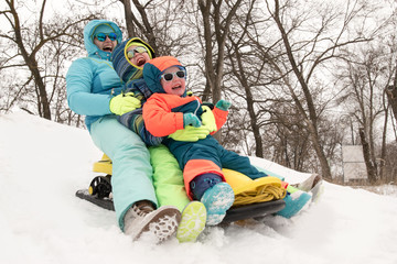 Happy mother with children in sunglasses and bright blue ski suit. Mother and boys are happy together. Kids ride a yellow sled in the forest with their mom. Guys are very happy to play outdoors