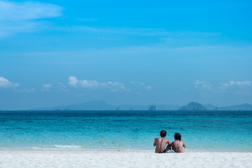 Young couple sitting by white sand together looking at the blue sea and sky.