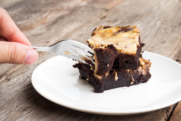Closeup woman using fork pick cheese brownies on white plate. Homemade dessert for chocolate lover. Served on wooden table.