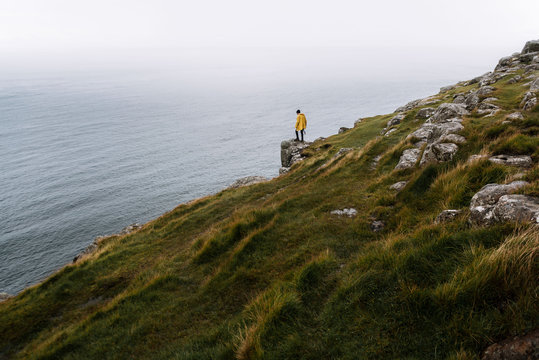 Young adult male wearing a yellow rain jacket standing on a overgrown cliff overlooking the overcast atlantic ocean in Scotland, UK