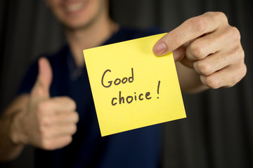 Man holds yellow sticker inscription Good choice! Thumb up Blue T-shirt. close-up, selective focus