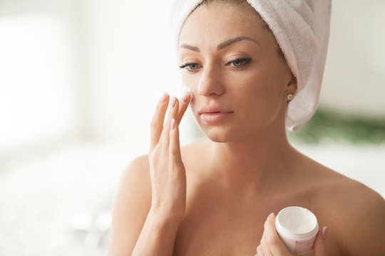 Young woman with flawless skin, applying moisturizing cream on her face. Photo of woman after bath in white bathrobe and towel. Skin care concept