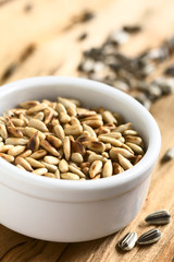 Roasted sunflower seeds in small bowl surrounded by whole seeds with hull, photographed on wood with natural light (Selective Focus, Focus in the middle of the seeds)
