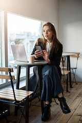 Can you bring receipt please. Indoor shot of stylish good-looking modern woman in local cafe sitting near window, drinking coffee while working in laptop, holding smartphone to call boss