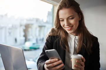 Come over to cafe in downtown. Portrait of happy stylish good-looking female student sitting in coffee shop, drinking beverage and messaging via smartphone, working on project with laptop