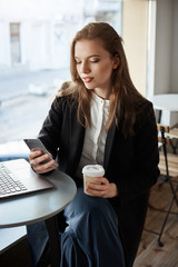 Excellent time for communication. Horizontal portrait of good-looking modern woman in cafe, sitting near window, drinking coffee and browsing in smartphone, having break of work with laptop